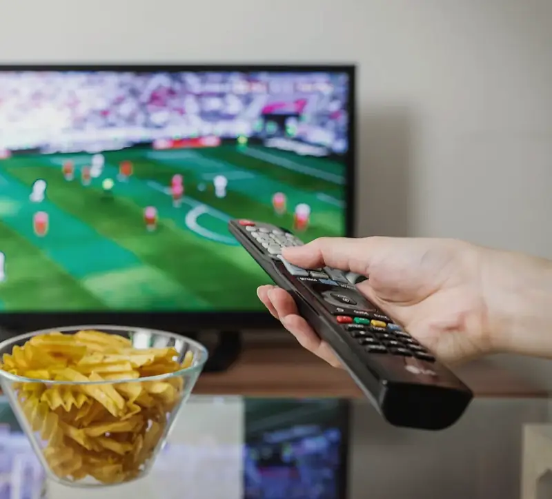 Match de football sur un téléviseur, une main tient la télécommande devant un bol de chips